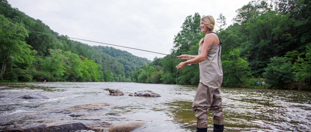 Woman fly-fishing in river surrounded by trees during daytime.