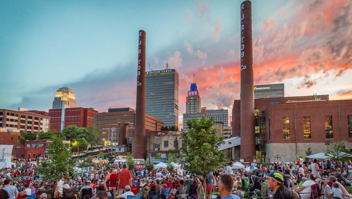 Large crowd of people sitting on lawn of park with Winston-Salem skyline in background
