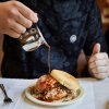 Person pouring barbecue sauce over pork sandwich on plate,