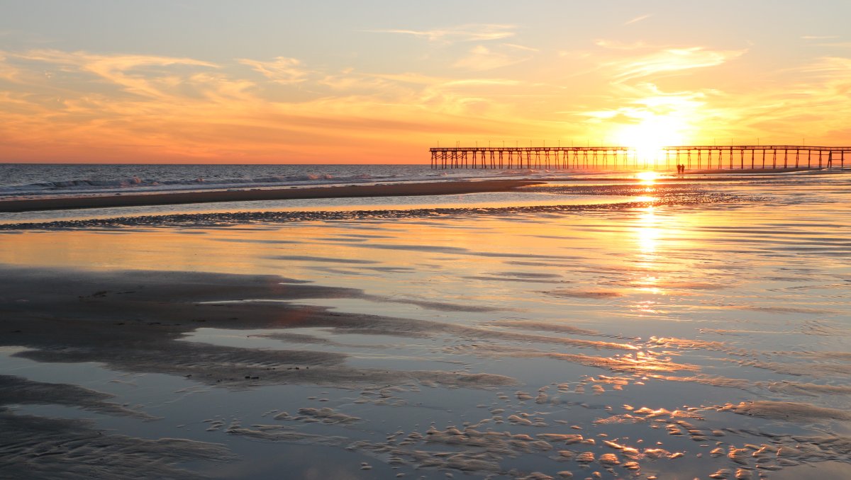 Long-range view of beach and ocean with sun setting behind pier in distance.