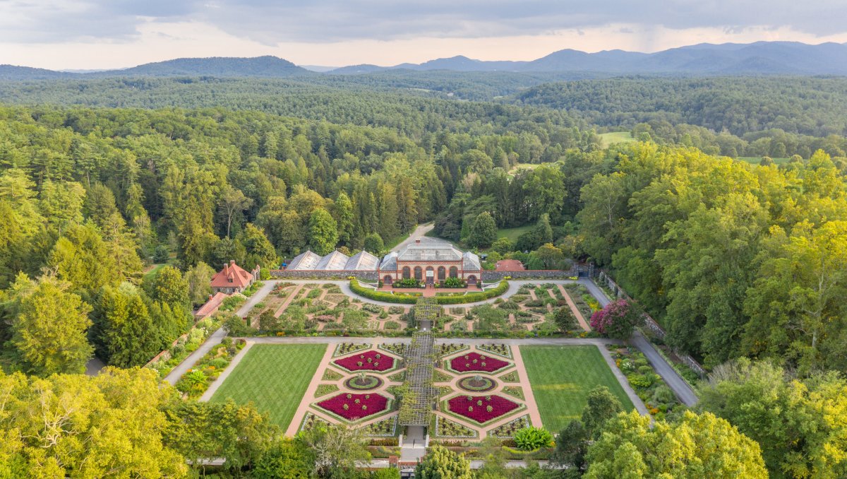  An aerial view of the meticulously designed Walled Garden at the Biltmore Estate, featuring vibrant geometric flower beds, manicured lawns, and lush greenery. The Conservatory stands at the garden’s center, framed by the surrounding forest and rolling Blue Ridge Mountains