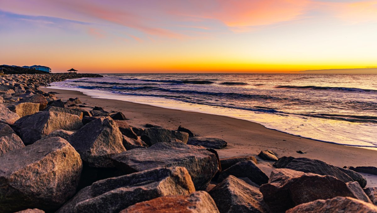 Sun rising over ocean on horizon with large rocks and sand in foreground