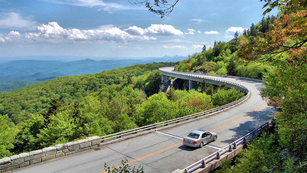Car driving on Linn Cove Viaduct on Blue Ridge Parkway with mountains in distance.
