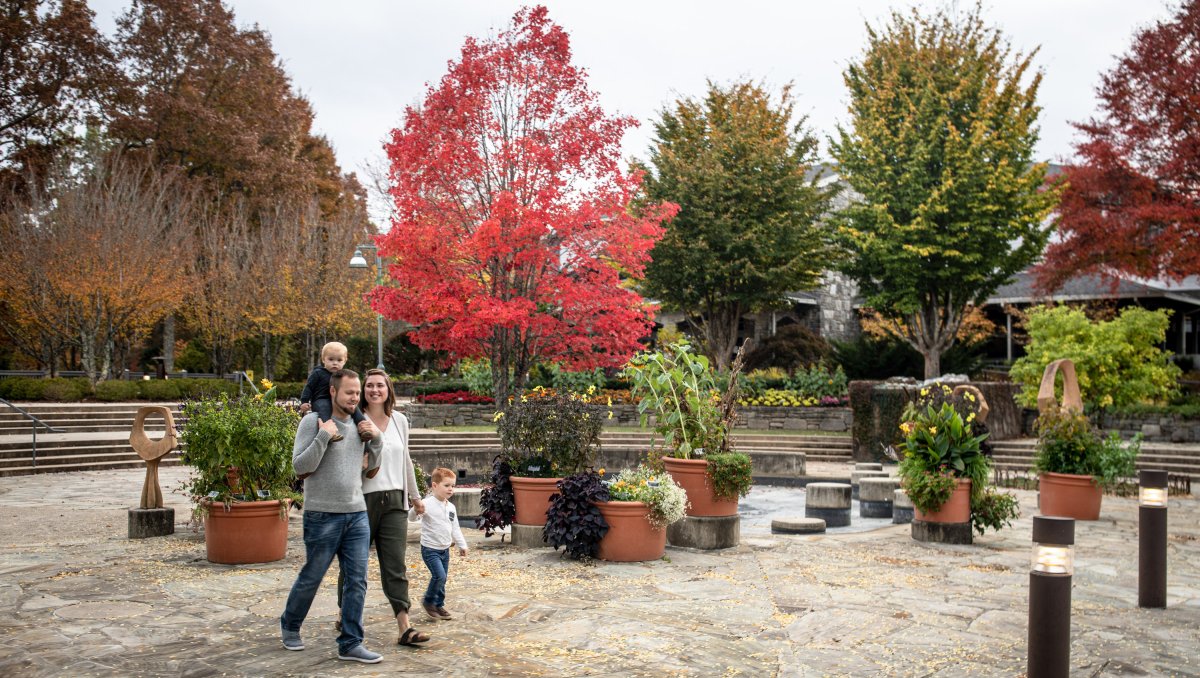 A family of four enjoys a stroll through a beautifully landscaped garden with colorful autumn trees in the background. The father carries a toddler on his shoulders, while the mother holds hands with a young child. Potted plants and stone pathways surround them, creating a warm and inviting atmosphere