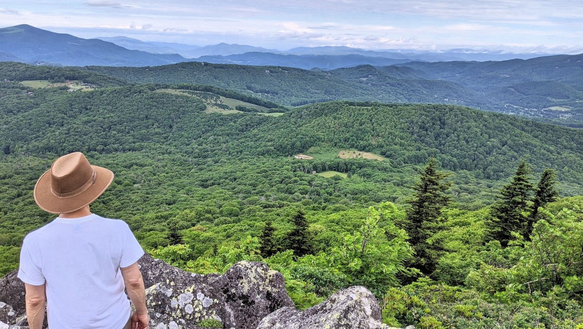 Person wearing hat standing at edge of cliff looking onto mountain range