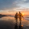 Backs of surfers walking in surf toward pier and sunrise.