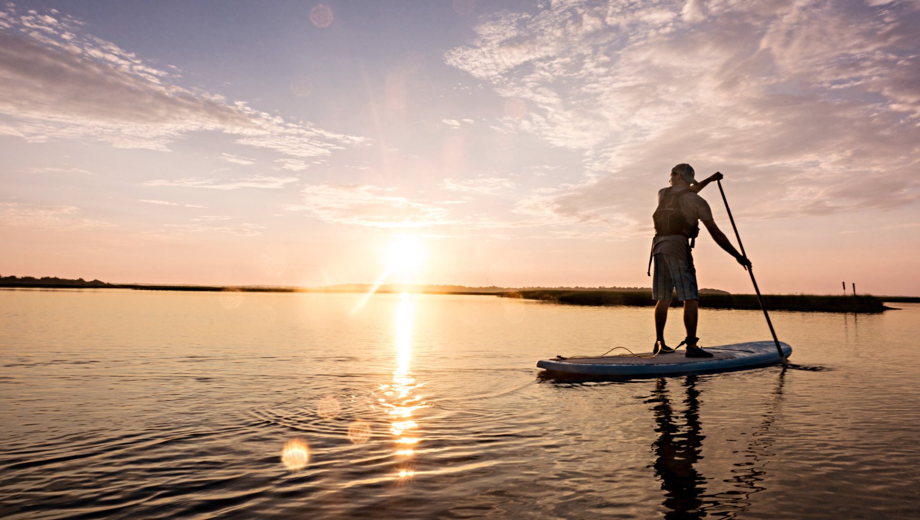 Person stand-up paddleboarding in Hammocks Beach State Park with sun rising in distance.