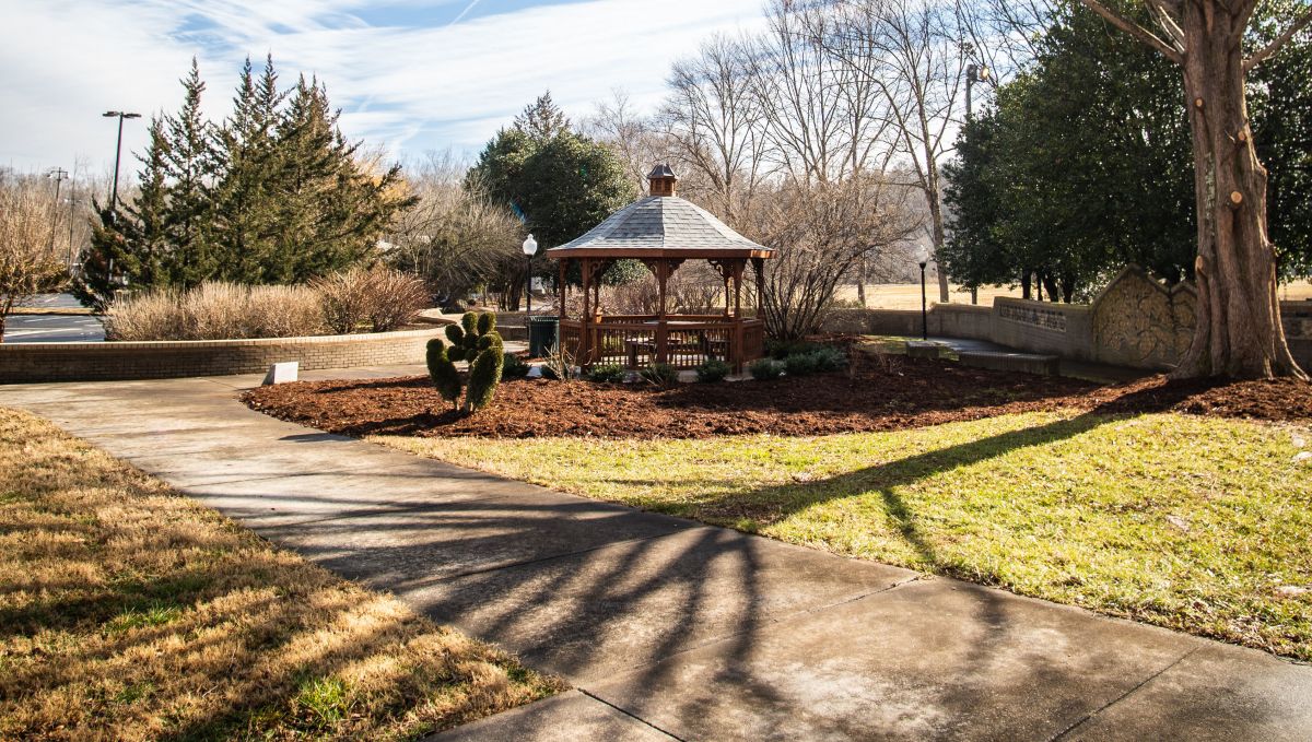 A serene outdoor scene featuring a wooden gazebo surrounded by winter landscaping. Bare tree branches cast intricate shadows on a paved pathway, while a mix of evergreens and dormant plants line the background. The sky is bright blue with wispy clouds, adding to the peaceful atmosphere.