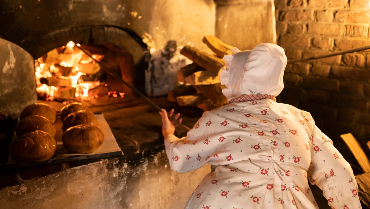 Back of Moravian baker inserting poker into wood fire at bakery